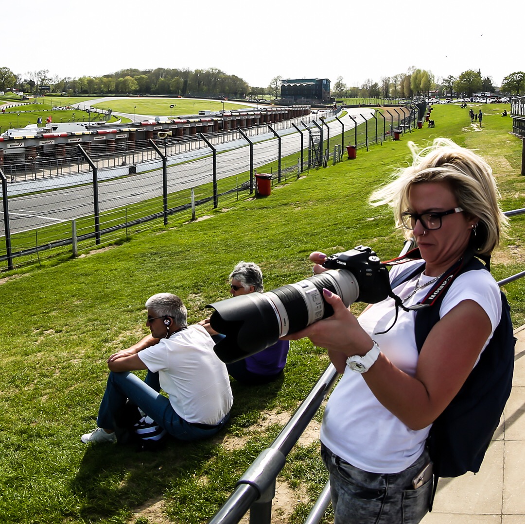 Natalia on the grandstand at Brands Hatch, telephoto lens in hand, race circuit sweeping behind.