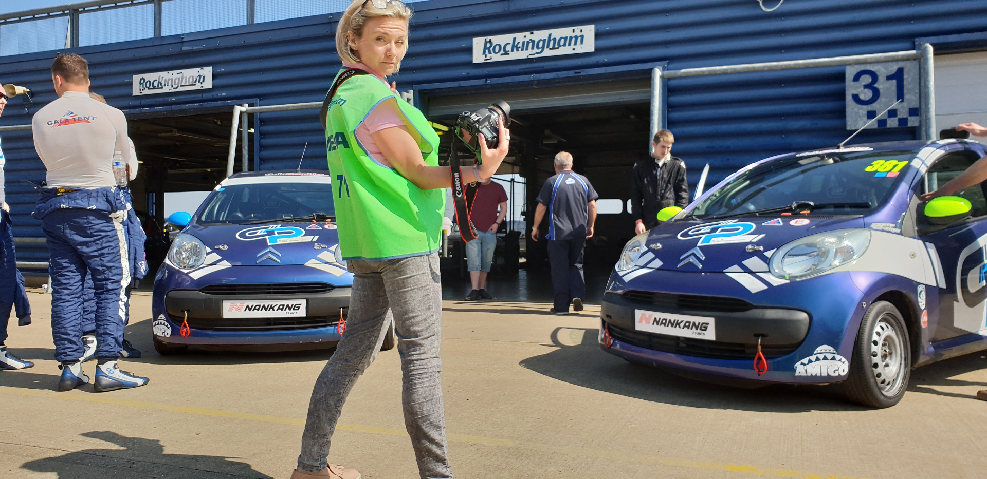 Natalia walking the pit lane in a hi-vis vest, camera in hand, race cars staged either side.