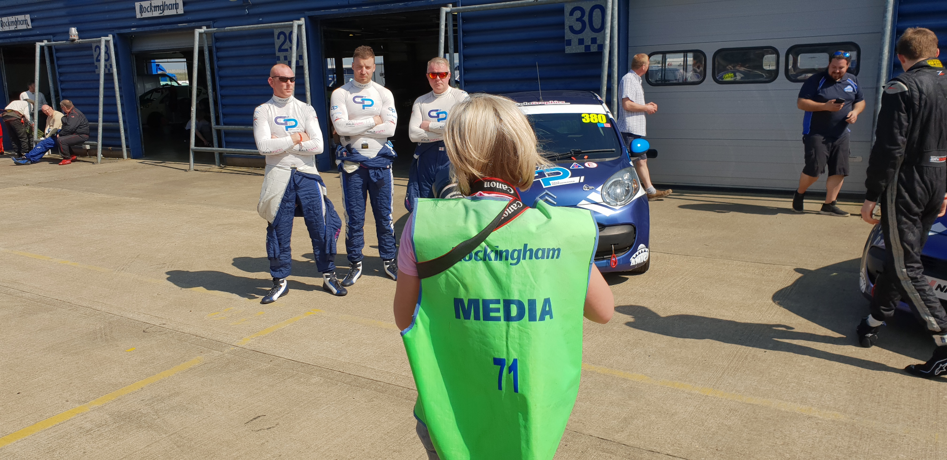 Natalia in the media vest, lens raised at three drivers leaning on the pit wall — the founder behind the frame.