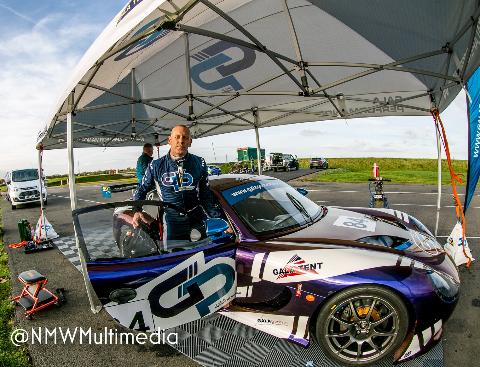 A driver suited up beside his Ginetta, paddock tent overhead, sky breaking through.